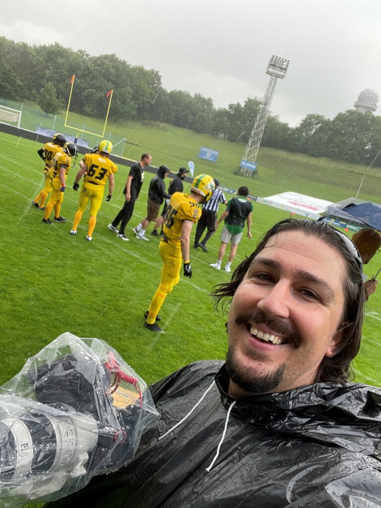 Sportvideograf Daniel Farkas mit Kamera in Plastiksack bei strömendem Regen am American Football Feld